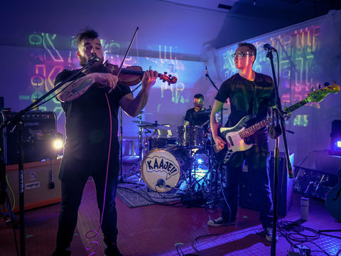 KAAJE!! band members performing on stage, one playing a guitar, one playing a violin, blue lights, green lights, pink lights are being reflected in the background