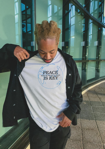 Man standing infront of windows wearing a 'Peace is key: Music is the key to peace' white t-shirt